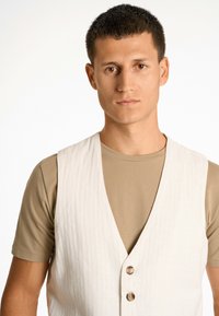 Weiße Leinenweste mit V-Ausschnitt-Design, ausgestattet mit zwei braunen Knöpfen. Getragen über einem enganliegenden beigen T-Shirt, glatte Textur, minimalistische Optik.