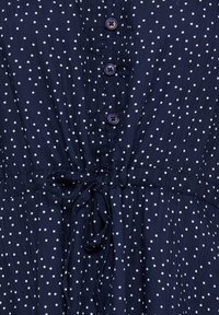 Tmavomodré šaty s bielymi bodkami, s štyrmi gombíkmi vpredu a látkovým opaskom v páse.