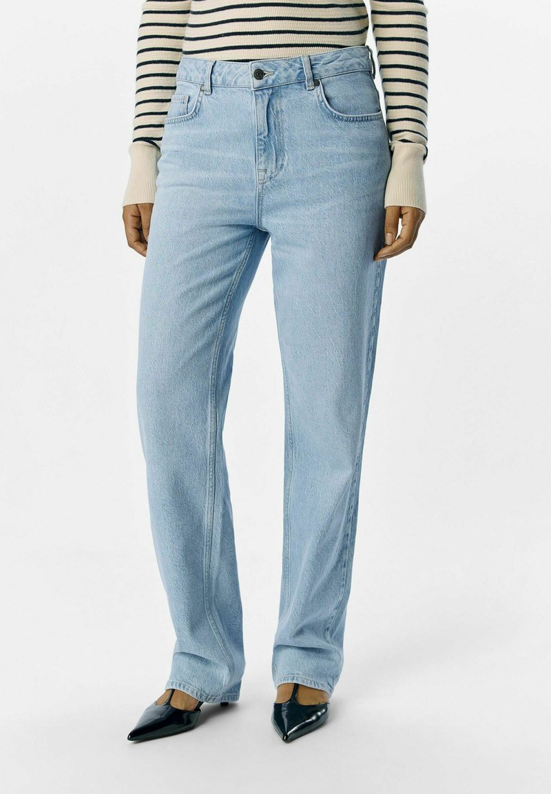 Svijetloplave traperice ravnog kroja, s dva prednja džepa i vidljivim dugmetom za zatvaranje. U kombinaciji s tamnozelenim cipelama šiljatog vrha.