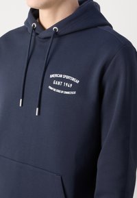 Marineblå hettegenser laget av mykt stoff, med lommer foran og snøring. Teksturert mønster med hvitt brodert logo og detaljer.