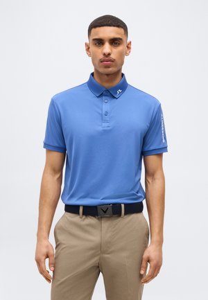 Άνδρας που φοράει μπλε κοντομάνικη πόλο μπλούζα με λογότυπο στο γιακά και το μανίκι, μπεζ παντελόνι και μαύρη ζώνη, στέκεται μπροστά σε λευκό φόντο.