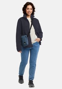 Navy fleece jacket, cream top, blue jeans, black shoes. Small blue crossbody bag with black logo, person smiling while walking.