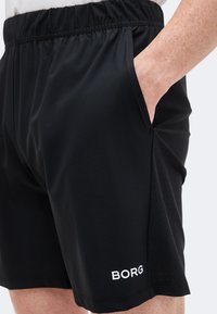 Κοντινό πλάνο μαύρων αθλητικών σορτς με ελαστική μέση, πλαϊνή τσέπη και το λευκό λογότυπο "BORG" στο κάτω μέρος του ποδιού, φορεμένα από άτομο με το χέρι στην τσέπη.