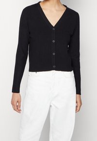 Cárdigan negro de canalé con mangas largas, escote en V y cierre de botones frontal. Llevado sobre jeans blancos, destacando los colores en contraste.