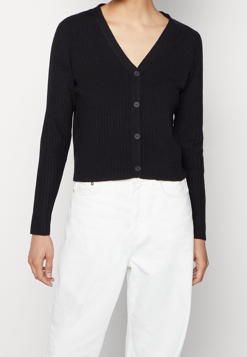 Cárdigan negro de canalé con mangas largas, escote en V y cierre de botones frontal. Llevado sobre jeans blancos, destacando los colores en contraste.