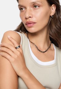 Ketting van schakels in donker metaal, ring in bijpassend ontwerp. Model draagt een lichtbeige tanktop met witte rand, heeft een gemiddelde huidtint en lichtbruin haar.