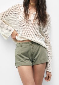 Jersey blanco de punto abierto con mangas largas y escote en V, combinado con pantalones cortos verde oliva con puños y cierre frontal de botón.