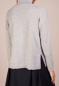 Pull à col roulé tricoté gris clair, manches longues, fentes latérales et texture douce, porté sur une jupe en tissu foncé.