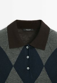 Harmaa ja laivastonsininen argyle-poolopaita ruskealla kauluksella, jossa on nappikiinnitys edessä sekä neljä nappia. Materiaali näyttää pehmeältä ja rakenteelliselta.