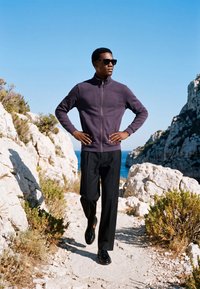 Homme portant des lunettes de soleil, une veste zippée sombre et un pantalon noir, debout sur un chemin rocheux avec la mer et des falaises en arrière-plan.