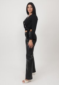 Top nero cropped a maniche lunghe con bottoni e dettagli confiati, abbinato a pantaloni neri in pelle ecologica aderenti e sandali con tacco a blocco di colore chiaro.