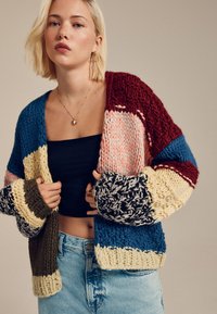 Chunky knit cardigan with multicolor stripes in burgundy, blue, cream, olive, and black, featuring oversized sleeves and textured patterns.