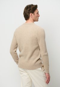 Beige ribbad tröja med långa ärmar och rund hals, med ett figurnära nederkant, visad bakifrån, tillsammans med ljusa byxor.