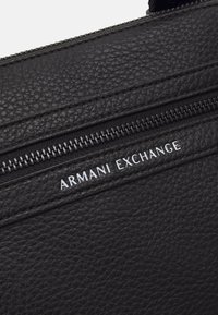 Borsa in pelle nera con texture, dotata di una cerniera, con il logo bianco "Armani Exchange" sul davanti. Il design è elegante e funzionale.