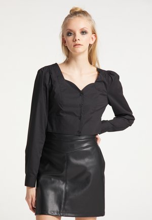 Junge Frau mit blondem zurückgebundenem Haar, die eine schwarze, geknöpfte Bluse mit Puffärmeln und einen schwarzen Leder-Minirock trägt.