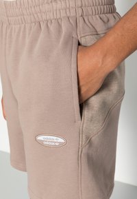 Bruna bomullsshorts med elastisk midja, sidofickor och en texturerad lapp. Har en broderad Adidas-logga vid fållen.
