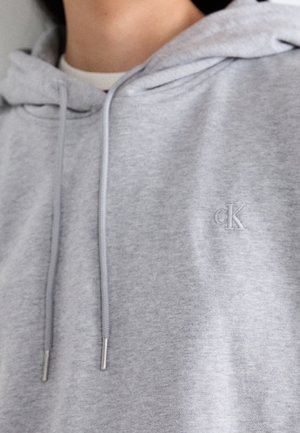Zbliżenie osoby noszącej jasnoszarą bluzę z kapturem Calvin Klein z troczkami i małe haftowane logo „cK” na piersi.
