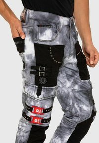 Pantalons en denim avec un motif tie-dye en gris et noir, présentant plusieurs poches, des éléments en métal, des clous et des boucles rouges pour une touche de détail.