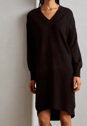 Kobieta ubrana w długą czarną sukienkę swetrową z dekoltem w serek i długimi rękawami, stojąca na tle gładkiej jasnoszarej ściany.