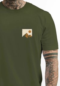 Olive green T-shirt with a small, centered pocket graphic featuring a sunset over mountains and trees, printed in beige and orange tones.