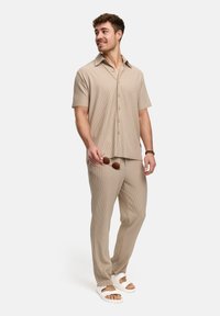 Beige, kortebladige knoopshirt met een ontspannen pasvorm, gecombineerd met bijpassende gestreepte broek. Sandalen en zonnebril in één hand.