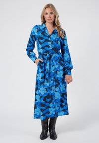 Niebieska sukienka w kwiaty z kołnierzykiem, długimi rękawami i guzikami z przodu. Posiada pasek nawiązujący w talii i kieszenie, osadzona na czarnym tle.