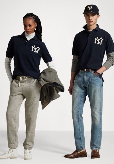 Marine polo med NY-logo, grå joggebukser og hvite sneakers. Mann i jeans og brune sko, holder en teksturert blazer. Visuell fokus på klærne.