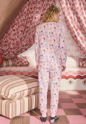 Nő hátulról fényképezve, rózsaszín pizsamát visel, amely színes kutya mintákkal díszített, egy virágos és csíkos mintájú szobában áll.