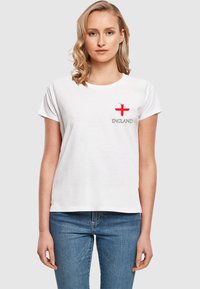 Balta medvilninė marškinėlė, kuriuose ant kairiojo krūtinės ploto esantis raudonas kryžius ir pilka spalva užrašyta „ENGLAND“. Trumpomis rankovėmis ir apvali iškirptė.