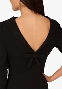 Czarna sukienka z głębokim dekoltem na plecach w kształcie litery V, ozdobiona kokardą w talii oraz o dopasowanym, strukturalnym kroju. Gładka faktura i dopasowana sylwetka.
