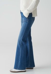 Uitlopende blauwe spijkerbroek met een medium wassing. Kenmerken zijn een hoge taille, zijzakken en een gladde textuur. Gecombineerd met witte sneakers.