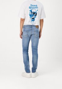 Valkoinen t-paita, jossa on sininen pääkallografiikka, yhdistetty vaaleansinisiin kapeisiin farkkuihin. Takana taskut ja hillitty farkkusaumaus. Valkoiset tennarit.