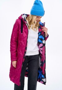 Dlouhý růžový prošívaný zimní kabát s květinovým vzorem na vnitřku, v kombinaci s bílým svetr a černými džíny. Modelka nosí modrou čepici.
