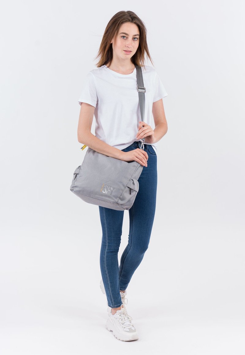 Mujer con camiseta blanca, jeans ajustados azules, zapatillas blancas y un bolso cruzado gris con una correa ancha sobre el hombro.