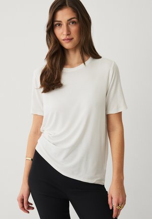 Kvinde med langt brunt hår iført en simpel hvid kortærmet trøje og sorte bukser, stående foran en neutral baggrund.