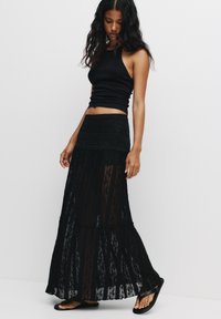 Jupe maxi en dentelle noire avec un design superposé et une taille en scallop, associée à un débardeur noir ajusté et des sandales plates noires.
