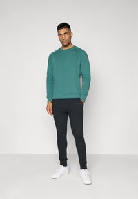 Teal lange mouwen sweatshirt met geribde manchetten en ronde hals, gecombineerd met zwarte tapered joggers en witte sneakers. Eenvoudig ontwerp, geen patronen.