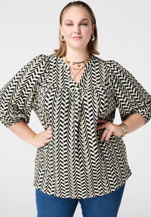 Vrouw draagt een zwarte, witte en beige blouse met zigzagpatroon en pofmouwen, blauwe jeans, gouden sieraden, poseert met handen in de zij.