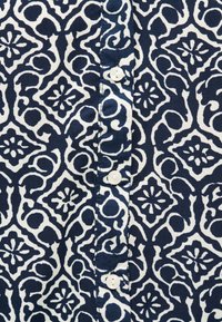 Tissu à fond blanc avec des motifs floraux et tourbillonnants bleus marine complexes, comportant une rangée verticale de boutons blancs.