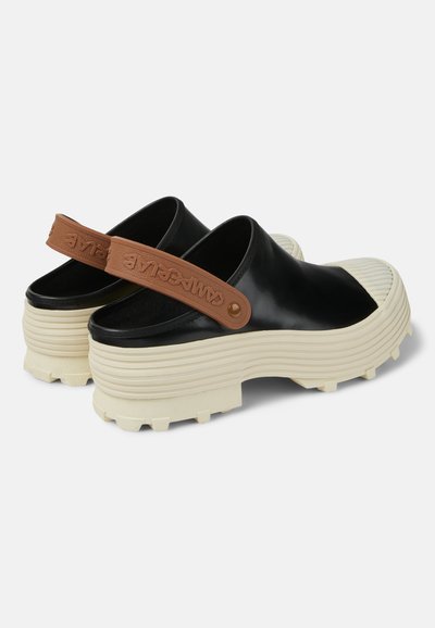 Svarta slip-on-skor med en texturerad gummisula, en knölig design och ett brunt justerbart band med präglad logotyp.