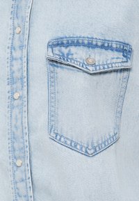 Light blue denim shirt featuring a pointed chest pocket with a silver snap button and structured stitching along the seams.