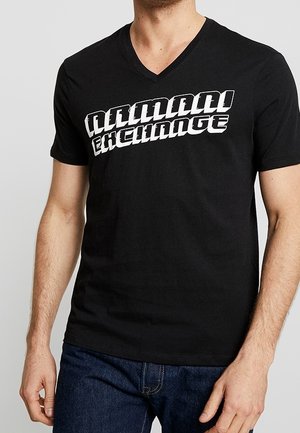 Mies, jolla on musta V-kaula-aukollinen T-paita jossa paksu valkoinen "Armani Exchange"-teksti, yhdistetty tummansinisiin farkkuihin, seisoo harmaan taustan edessä.