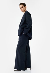 Marineblauwe pinstripe tailored blazer met bijpassende wijde broek. De stof lijkt glad met een subtiele glans. Opvallende oorbelaccenten.