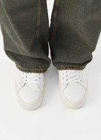 Λευκά δερμάτινα sneakers με επίπεδα κορδόνια και καουτσούκ σόλες, τοποθετημένα με σκούρο, ελαφρώς φθαρμένο τζιν παντελόνι που δείχνει λεπτομέρειες με ξέφτια στο στρίφωμα.