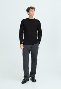 Svart ribbad stickad tröja med rundad halsringning, tillsammans med mörkgrå raka byxor och svarta skor, mot en ljus bakgrund.