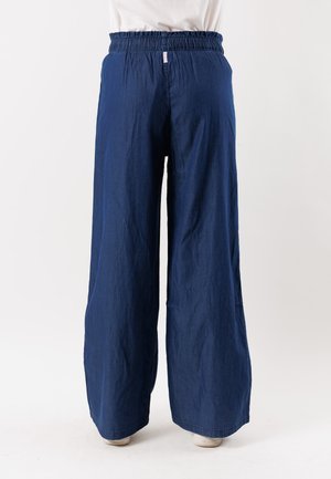 Marineblauwe broek met wijde pijpen en elastische tailleband, gedragen door een persoon die tegen een witte achtergrond staat.