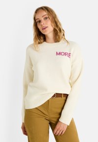 Pull en tricot crème avec des poignets et un ourlet côtelés, affichant le mot "MORE" en lettres roses sur la poitrine. Associé à un pantalon marron avec ceinture.