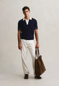 Férfi áll, sötétkék rövid ujjú pólót, fehér nadrágot, sötét cipőt visel, kezében nagy barna táskát tart, egyszerű világos háttér előtt.
