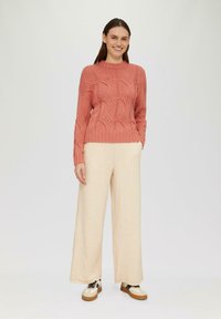 Koraal geknoopte trui met textuurpatroon, gecombineerd met crèmekleurige wijde broek. Schoenen zijn beige met zwarte en groene accenten.