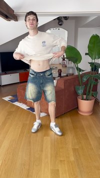 Beige pitkähihaisella paidalla ja kulutetuilla farkkushortsilla, jossa on sininen vyötärö. Hopeiset lenkkarit. Sisätila, jossa on kasveja ja huonekaluja.